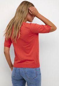 Red short-sleeve sweater with a fitted design and ribbed cuffs. Worn with light blue jeans, showing a smooth texture and casual style.
