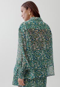 Floral patterned, sheer green blouse with long sleeves, a collared neck, and gathered cuffs. Features a lightweight, flowy fabric.