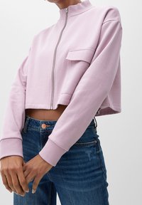 Maglione cropped viola chiaro con colletto alto, zip frontale e taschino singolo sul petto, abbinato a jeans blu con dettagli dei bottoni visibili.