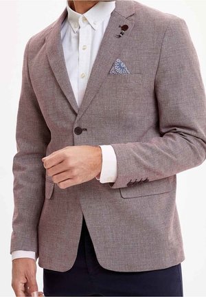 Hombre con blazer marrón claro texturizado, camisa blanca, pañuelo de bolsillo estampado y pantalones oscuros, ajustándose la chaqueta con la mano izquierda.