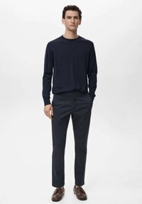 Navy-blaues Strickpullover mit Rundhalsausschnitt, kombiniert mit dunkelgrauen Hosen und braunen Lederschuhen, getragen von einem Modell vor einem neutralen Hintergrund.
