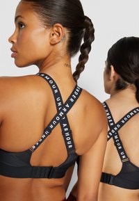 Černé sportovní podprsenky s křížovými ramínky a logem "UNDER ARMOUR"; nastavitelná zapínání na zadní straně; hladká textilie.