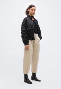 Νεαρή γυναίκα που φοράει μαύρο μπουφάν bomber, μαύρο τοπ, μπεζ παντελόνι με φαρδιά μπατζάκια και ψηλή μέση, μαύρα μποτάκια αστραγάλου και μαύρο μαντήλι με σχέδια στον λαιμό.