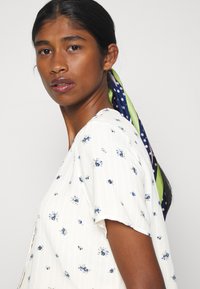 Blusa branca com pequenos padrões florais azuis, mangas curtas e decote em V. Cabelo preso com um lenço colorido com bolinhas e riscas.