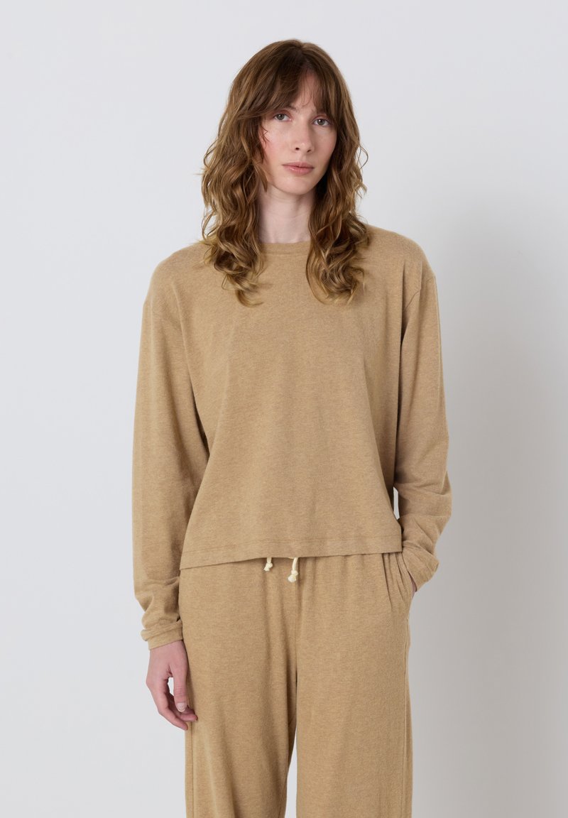 Jeune femme aux cheveux châtain clair ondulés portant un pull beige ample et un pantalon assorti à cordon de serrage, se tenant devant un fond gris uni.