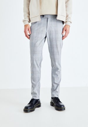 Muž ve béžovém saku, kostkovaných světle šedých slim fit kalhotách a černých kožených šněrovacích botách stojí na bílém pozadí.