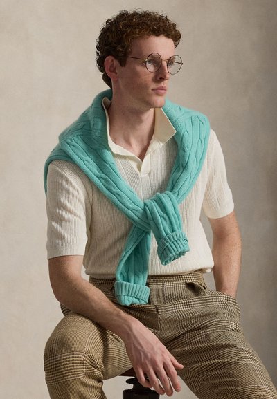 Νέος άνδρας με σγουρά μαλλιά και στρογγυλά γυαλιά, φορώντας λευκό πλεκτό μπλουζάκι με κοντό μανίκι και μπλε πράσινη ζακέτα περασμένη πάνω από τους ώμους, καθιστός.