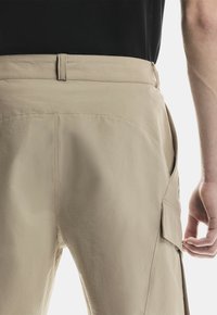 Pantalones cargo beige con solapa en el bolsillo lateral, llevados por una persona con camiseta negra, mostrados desde la parte baja de la espalda hasta la mitad del muslo.