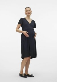 Marineblaffes Umstandskleid mit V-Ausschnitt, kurzen Ärmeln und geraffter Taille. Weicher, glatter Stoff, knielang mit lockerer Passform. Schwarze Ballerinas.