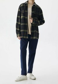 Camisa de franela a cuadros en verde oscuro y azul marino, abotonada, con dos bolsillos frontales; combinada con una camiseta beige, pantalones azul marino y zapatillas blancas.