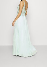 Mintově zelené večerní šaty s volánkovou sukní, korzetem zdobeným korálky a nízkým designem na zádech. Disponují tenkými ramínky a měkkou, texturovanou tkaninou.