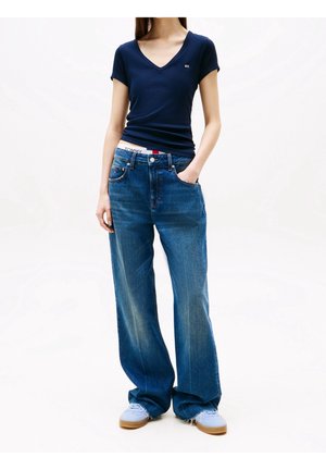 Vrouw draagt een marineblauw aansluitend T-shirt met V-hals, losse blauwe jeans, zichtbare witte tailleband en lichtblauwe sneakers, staand met één hand in de zak.