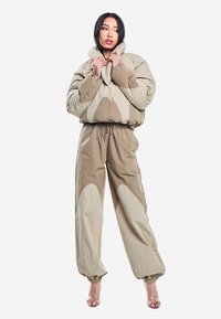 Beżowa kurtka puchowa z wysokim kołnierzem i abstrakcyjnym wzorem, zestawiona ze spodniami z elastyczną talią, ozdobionymi dużymi beżowymi i brązowymi akcentami.