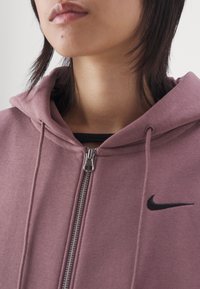 Hoodie i mjuk mauve-tyg med full dragkedja och en svart Nike-logotyp på bröstet. Närbild av halsområdet och huvan.