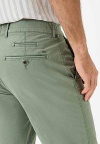 Pantaloni verde chiaro realizzati in materiale morbido, con tasche posteriori dotate di cuciture orizzontali e chiusura con bottone, contrassegnati da un piccolo logo.