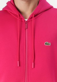 Fuchsieová mikina na zips s kengurím vreckom, nastaviteľnými šnúrkami a malým zeleným vyšitým logom krokodíla na hrudi.
