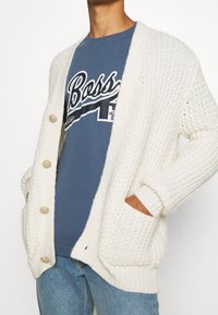 Vaalean beigen neulejakun, jossa on paksu rakenne, suuret napit, ribbineulehihat ja sivutaskut, käytetään sinisen graafisen t-paidan päällä.