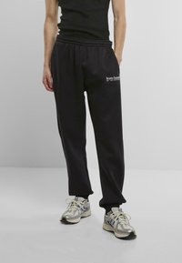 Black jogger pants with elastic waistband, side pockets, and white text logo. Paired with silver running shoes, textured finish.