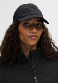 Čierna baseballová čiapka vyrobená z látky, s hladkým povrchom, s mierne zakriveným šiltom. Nosená s čiernou bundou na zips, ktorá má detaily šitia.