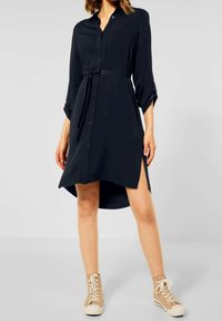 Svart knälång klänning med krage och bindbart midjeband, som har långa ärmar med uppvikt fåll, högt lågt fåll och sidoflikar. I kombination med beige sneakers.