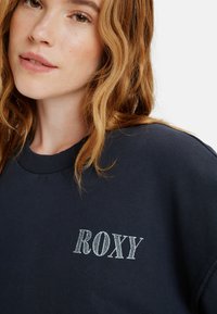 Marineblauwe sweatshirt met geribde halslijn en wit "ROXY" logo. Zachte textuur, casual pasvorm en minimale details.