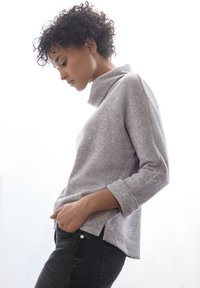 Grauer Pullover mit hohem Kragen und langen Ärmeln, aus einem weichen, strukturierten Stoff. Schwarze enge Jeans mit Seitentaschen. Modell posiert seitlich.