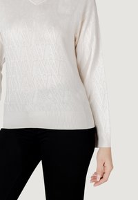 Maglione a maglia color beige chiaro con motivo a diamante testurizzato, scollo a V, maniche lunghe e orlo a coste. Abbinato a pantaloni neri attillati.