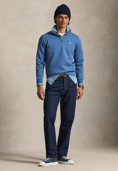 Ανοιχτό μπλε φλις μπλουζάκι με φερμουάρ μέχρι τη μέση, φορεμένο πάνω από ανοιχτό τζιν πουκάμισο, συνδυασμένο με σκούρα τζιν και μπλε αθλητικά παπούτσια.