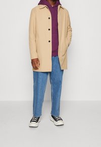 Uomo con un cappotto beige abbottonato sopra un felpa viola, jeans blu e sneakers bianco e nere, in piedi davanti a uno sfondo chiaro uniforme.