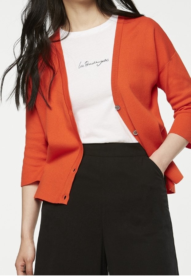 Orange kofta med trekvartslånga ärmar, utrustad med två knappar och en strukturerad stickning. Bärs över en vit T-shirt med svart text och svarta byxor.