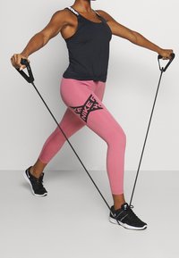 Débardeur de sport noir avec un nœud à l'avant, leggings roses avec un logo noir, et chaussures de sport noires. Bandes de résistance utilisées.