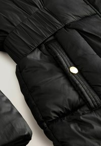 Doudoune noire à texture brillante, avec un design matelassé, une taille élastique et un accent de bouton-pression doré.