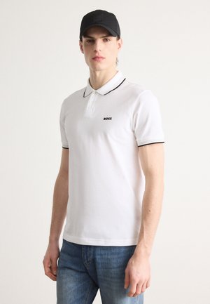 Polo alb cu guler negru și logo brodat. Purtați cu blugi albaștri, modelul poartă o șapcă neagră. Potrivire casual și textură netedă.