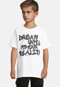 Balta medvilnė marškinėliai su ryškiu juodu grafiti tekstu "SVAJOK, KOL TAI TAPO TAVO REALYBE." Trumpomis rankovėmis ir apvaliu kaklu.