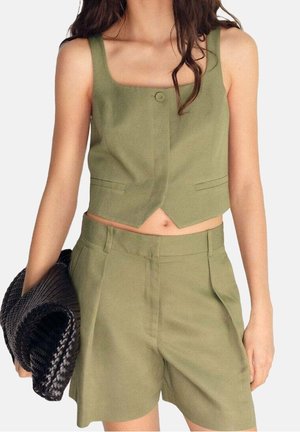 Femme portant un gilet court sans manches vert olive et un short taille haute assorti, tenant une grande pochette tissée noire.