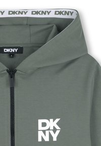 Sweat à capuche gris zippé avec une texture douce, arborant un logo DKNY blanc sur la poitrine gauche et une bordure intérieure de capuche blanche contrastante.