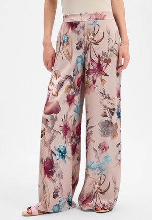 Pantalon large rose clair avec un grand imprimé floral rouge, bleu et beige, porté avec des sandales ouvertes et un haut beige.