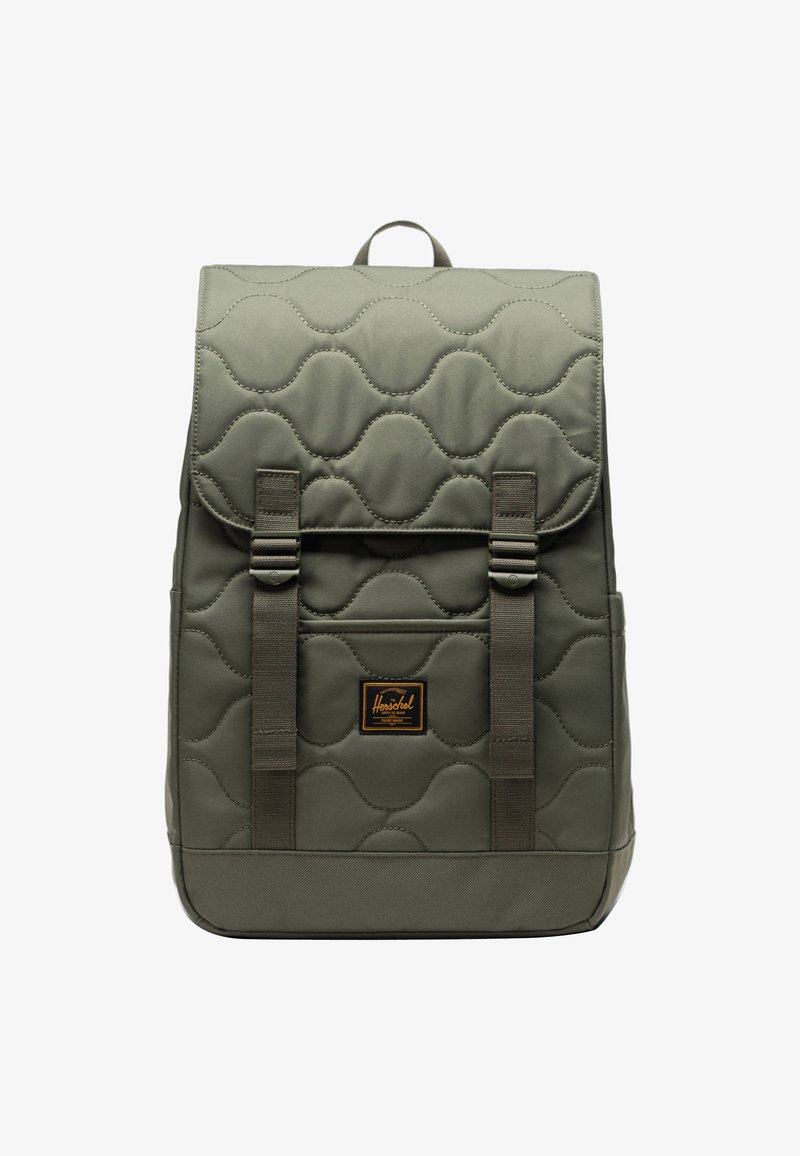 Zaino verde trapuntato con un pattino quadrato, due cinturini frontali, una maniglia superiore e una toppa con logo. Presenta superfici lisce e testurizzate.