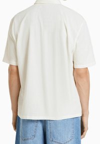 Camicia bianca a maniche corte con finitura texture, dotata di colletto e vestibilità rilassata. Il design sul retro è semplice, con sottili linee verticali.