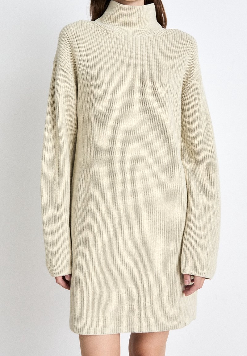 Femme portant une robe pull en maille côtelée beige à col roulé et manches longues, montrée des épaules jusqu'au milieu des cuisses sur fond blanc.