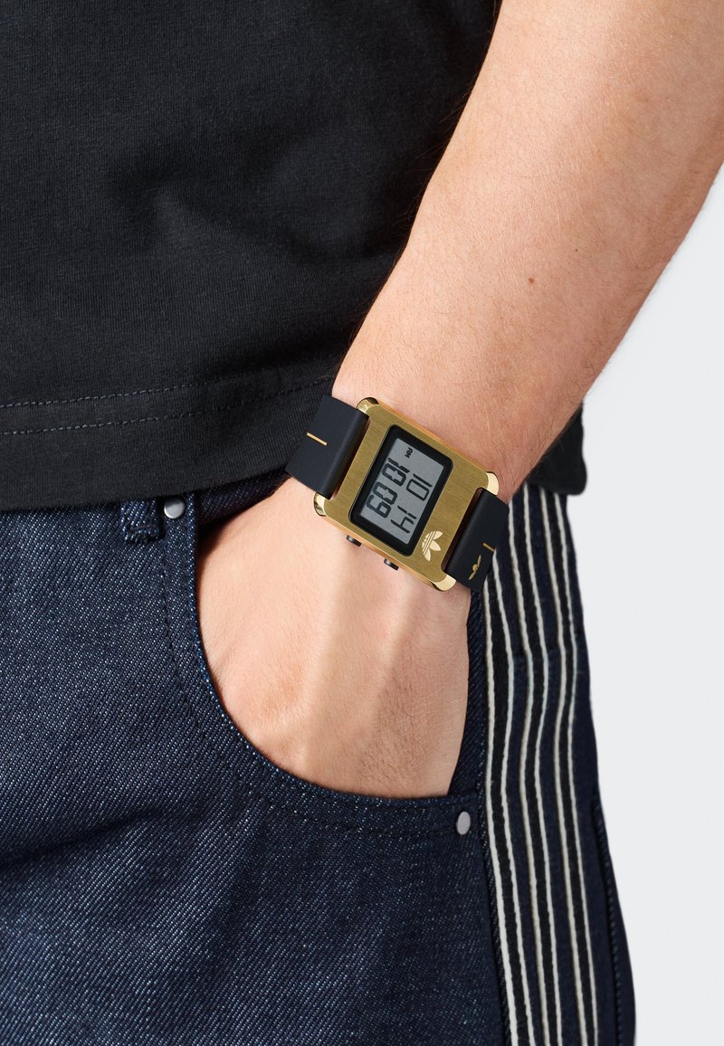 Roka zlate digitalne ročne ure z črnim trakom, palec ujet v žep temno modrih prugastih hlačah, vidna črna majica.