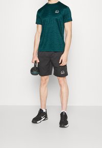 Zielona sportowa koszulka w wzory, czarne spodenki i czarne trampki. Trzyma czarny kettlebell z niebieskimi akcentami. Minimalne oznaczenia marki.