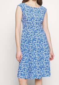 Niebieska sukienka w kwiatowy wzór z krótkimi rękawami, dopasowanym górą i rozkloszowanym dołem. Materiał jest lekki, o miękkiej teksturze i detalach w postaci fałd.