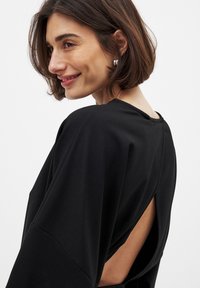 Schwarzes Kleid mit offener Rückseite, ausgestattet mit fließenden Ärmeln und einer glatten Textur. Akzente sind ein runder Ausschnitt und elegante Drapierungen.