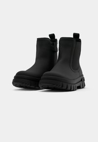Bottines noires avec un matériau lisse, semblable à du cuir, des panneaux latéraux élastiques et une semelle épaisse et texturée en caoutchouc. Elles sont dotées de languettes pour un enfilage facile.