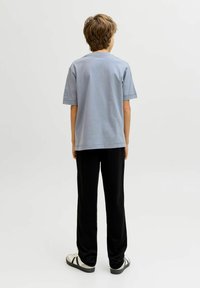 Νεαρό άτομο που στέκεται με την πλάτη, φοράει ένα ανοιχτό γαλάζιο μπλουζάκι, μαύρο παντελόνι και λευκά αθλητικά παπούτσια με μαύρες ρίγες, σε απλό ανοιχτό φόντο.