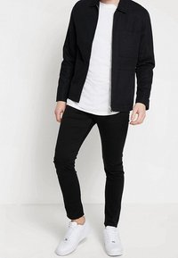 Chaqueta negra con cremallera y un bolsillo frontal, llevada sobre una camiseta blanca y pantalones ajustados negros, combinada con zapatillas blancas.