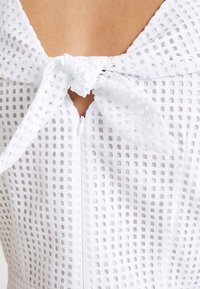 Vestido blanco texturizado con patrón de recortes cuadrados, con un lazo atado en el escote y cierre de cremallera en la parte delantera.