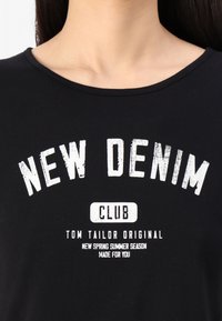 Melnas T-krekls ar noapaļotu kakla izgriezumu, uz kura ir balti drukāti teksti: "JAUNĀ DENIM KLUBS" un "TOM TAILOR ORIĢINĀLS" dažādos izmēros.