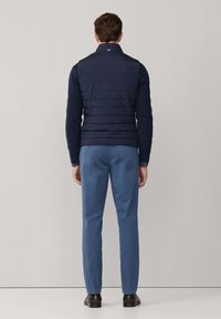 Marineblaue gesteppte Weste über einem strukturierten Pullover, kombiniert mit hellblauen Hosen. Das Ensemble zeichnet sich durch einen Stehkragen und glattes Material aus.